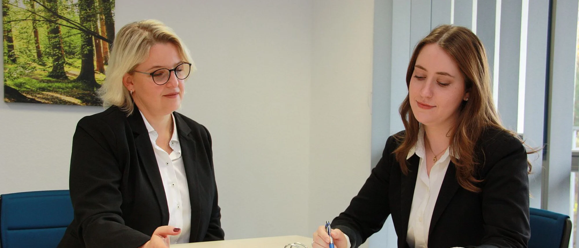 Das Bild zeigt zwei Frauen in professioneller Kleidung, die an einem Tisch sitzen und an einem Ordner mit der Aufschrift „Qualitätssicherungsrichtlinien“ arbeiten. Eine Frau liest aufmerksam ein Dokument, während die andere Notizen macht. Der Raum wirkt sachlich und ordentlich, mit dezenten Dekorationen wie einem Bild eines Waldes im Hintergrund, was eine ruhige und konzentrierte Arbeitsatmosphäre schafft.