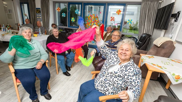 Auf dem Bild sind mehrere ältere Personen zu sehen, die bunte Tücher in der Hand halten und sie fröhlich schwingen. Die Szene wirkt lebhaft und zeigt eine Gruppeaktivität in einem Gemeinschaftsraum mit dekorierten Fenstern im Hintergrund. Die Stimmung ist ausgelassen, und die Teilnehmer scheinen Spaß an der Bewegung zu haben.