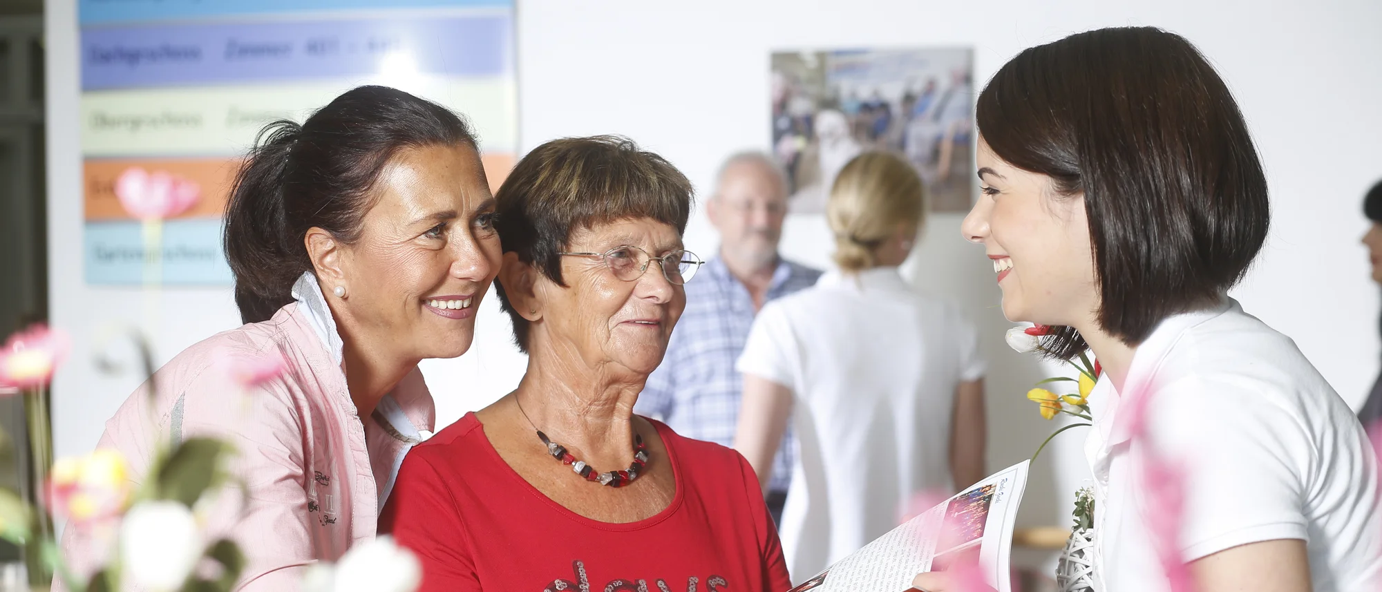 Das Bild zeigt eine fröhliche Interaktion zwischen drei Frauen. Eine ältere Frau in einem roten Shirt mit einer auffälligen Halskette hält ein Heft, das von einer jüngeren Frau in weißer Kleidung präsentiert wird. Neben der älteren Frau steht eine weitere Frau in rosa Kleidung, die sie freundlich anlächelt. Der Hintergrund ist lebendig mit unscharfen Personen und dekorativen Elementen, die eine angenehme, einladende Atmosphäre schaffen. Im Vordergrund sind Blumenarrangements sichtbar, die das Bild farbenfroh ergänzen.