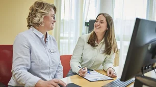Das Bild zeigt zwei Frauen, die an einem Schreibtisch sitzen und sich in einer freundlichen und konzentrierten Atmosphäre unterhalten. Beide tragen helle Blusen und Namensschilder, was auf eine professionelle Umgebung hinweist. Vor ihnen stehen ein Computer und ein Klemmbrett mit Dokumenten, was auf eine gemeinsame Arbeits- oder Schulungssituation hindeutet. Das einfallende Tageslicht und die hellen Farben des Raumes sorgen für eine angenehme Atmosphäre.