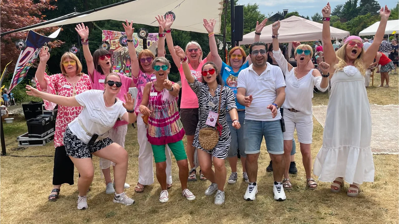 Eine fröhliche Gruppe von Menschen steht draußen bei einem Sommerfest und posiert lachend und jubelnd für ein Foto. Viele der Personen tragen farbenfrohe Kleidung, Sonnenbrillen und Accessoires, was eine ausgelassene und sommerliche Stimmung vermittelt. Im Hintergrund sind Bäume, Zelte und Dekorationen zu sehen, die die Festatmosphäre unterstreichen.