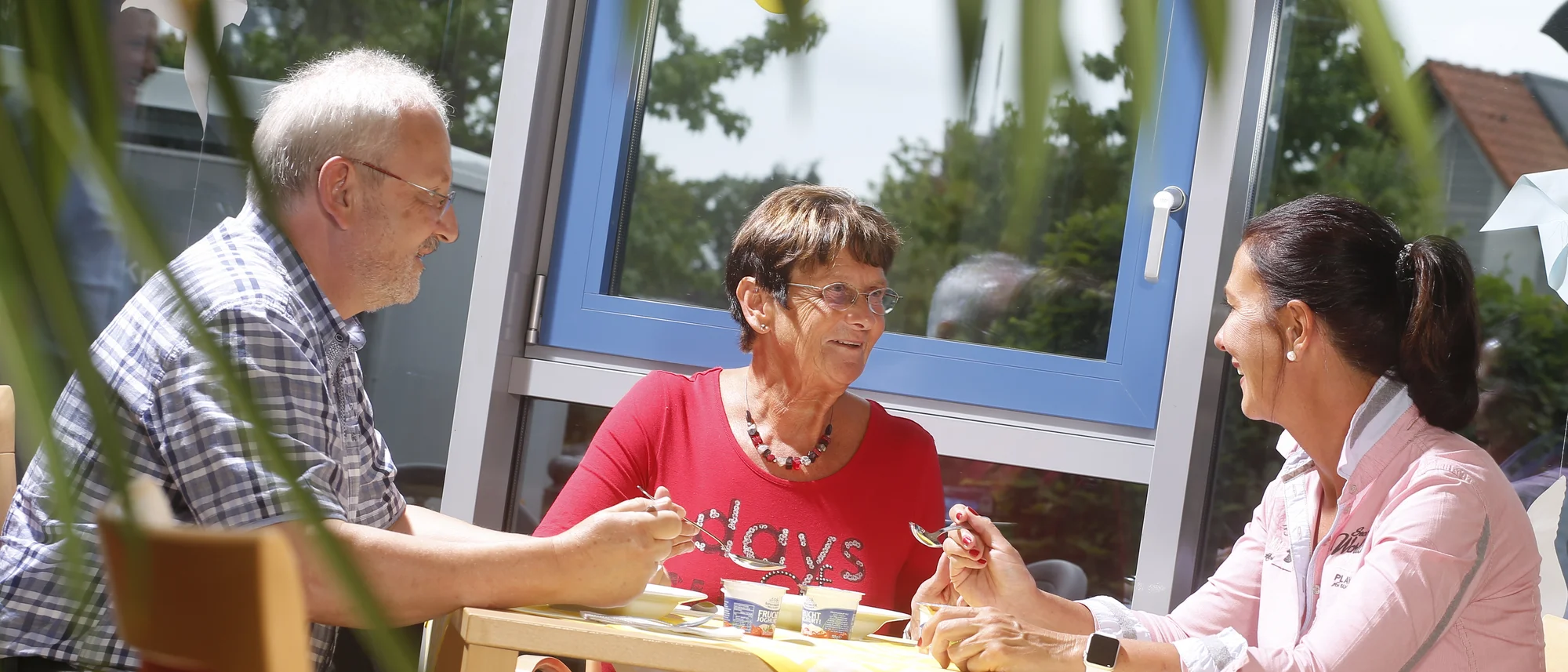 Das Bild zeigt drei Personen, die an einem Tisch in einer hellen und freundlichen Umgebung sitzen. Eine Frau im roten Shirt lächelt und unterhält sich mit einem Mann in kariertem Hemd und einer Frau in rosa Bluse. Auf dem Tisch liegen gelbe Tischdecken mit weißem Muster, kleine Joghurtbecher und Besteck. Im Hintergrund sind ein Fenster mit Blick ins Grüne und Dekorationen wie ein Windrad sichtbar. Die Szene strahlt eine entspannte und gesellige Atmosphäre aus.