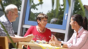 Das Bild zeigt drei Personen, die an einem Tisch in einer hellen und freundlichen Umgebung sitzen. Eine Frau im roten Shirt lächelt und unterhält sich mit einem Mann in kariertem Hemd und einer Frau in rosa Bluse. Auf dem Tisch liegen gelbe Tischdecken mit weißem Muster, kleine Joghurtbecher und Besteck. Im Hintergrund sind ein Fenster mit Blick ins Grüne und Dekorationen wie ein Windrad sichtbar. Die Szene strahlt eine entspannte und gesellige Atmosphäre aus.