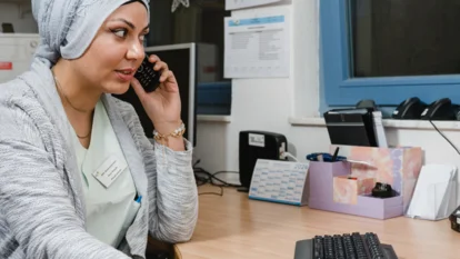 Eine Frau mit hellgrauem Kopftuch und heller Strickjacke sitzt an einem Schreibtisch und telefoniert. Mit einer Hand hält sie das Telefon ans Ohr, mit der anderen bedient sie eine Tastatur. Im Hintergrund sind ein Kalender, ein Fenster und verschiedene Büromaterialien zu sehen.