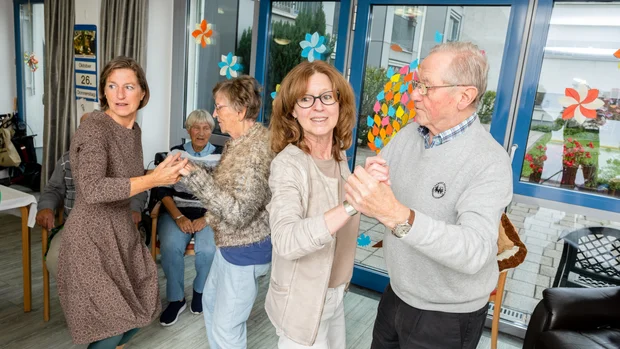 Auf dem Bild tanzen zwei Paare fröhlich in einem Raum. Die Personen sind älter und wirken entspannt und gut gelaunt. Im Hintergrund sitzen weitere Menschen und beobachten die Szene, während die Fenster mit bunten Papierblumen dekoriert sind. Die Atmosphäre ist gemütlich und zeigt ein geselliges Beisammensein.