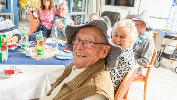 Auf dem Bild ist ein älterer Mann mit Brille und einem grauen Filzhut zu sehen, der fröhlich in die Kamera lächelt. Neben ihm sitzt eine ältere Dame, die ebenfalls lächelt. Im Hintergrund sitzen weitere Personen an einem geschmückten Tisch, der mit bayerischen Motiven dekoriert ist. Die Szene vermittelt Freude und Gemeinschaft bei einem Fest.