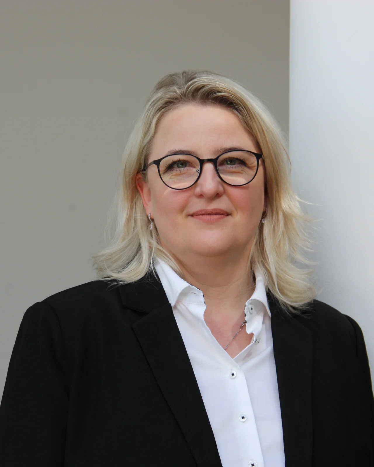 Eine Frau mit mittellangem, blonden Haar in einem schlichten schwarzen Blazer. Sie hat ein freundliches Lächeln und steht vor einem hellen Hintergrund