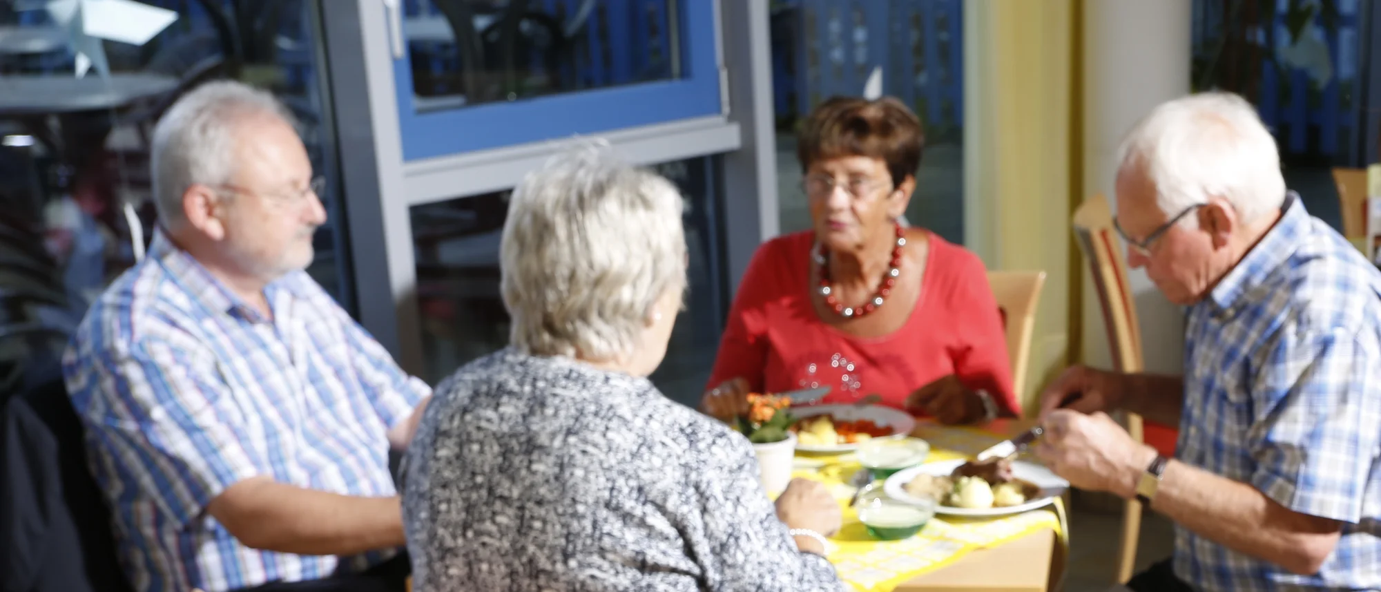 Eine Frau trägt ein Tablett mit zwei schön angerichteten Tellern, auf denen ein Fleischgericht mit Kartoffeln und Kraut sowie ein bunter Salat mit Mais und Gemüse zu sehen sind. Im Hintergrund sitzen vier ältere Menschen an einem Tisch und genießen gemeinsam ihr Essen, während sie sich unterhalten. Der Raum wirkt hell und freundlich durch große Fenster, die viel Tageslicht hereinlassen.