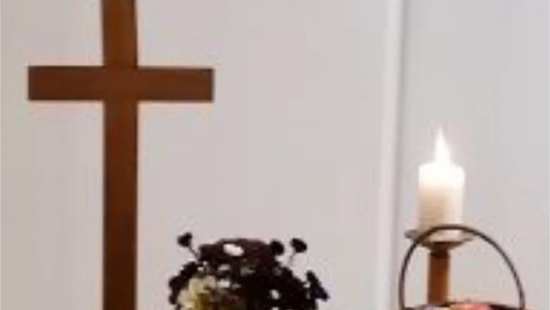 Auf einem Tisch steht ein schlichtes hölzernes Kreuz, daneben eine brennende weiße Kerze auf einem Holzständer. Ein kleiner Korb mit Früchten, darunter ein Apfel, und ein Blumenarrangement ergänzen die Dekoration. Der Hintergrund ist neutral, was den Fokus auf die symbolische Gestaltung des Altars lenkt.