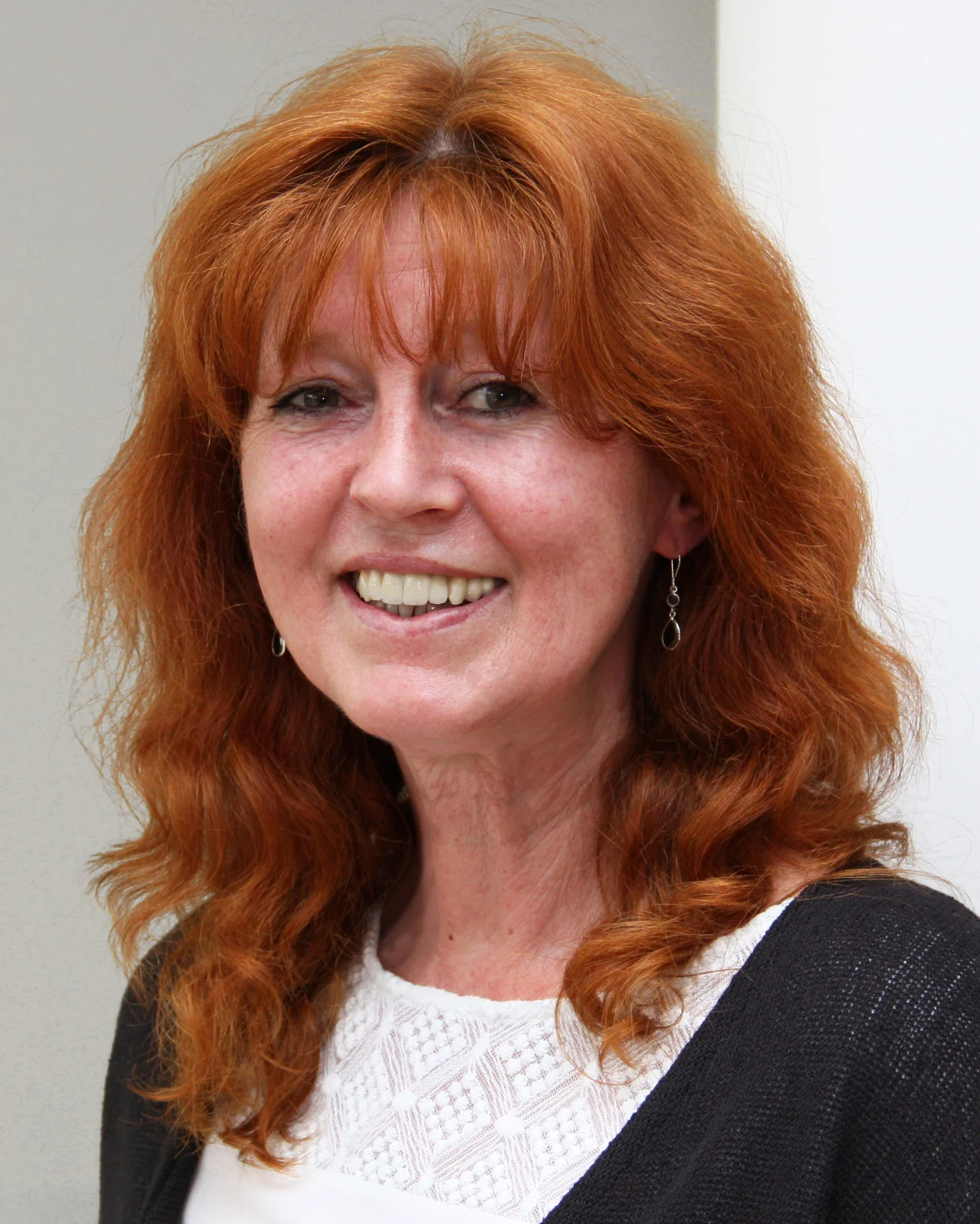 Eine Frau mit langen roten Haaren und einem herzlichen Lächeln ist zu sehen. Sie trägt eine weiße Bluse und einen schwarzen Cardigan.