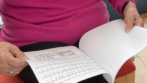 Eine Person in einem pinken Pullover blättert in einem Liederbuch, das Noten und Texte eines Liedes zeigt. Auf der aufgeschlagenen Seite sind Notenzeilen und ein kleiner schwarz-weißer Druck zu sehen. Das Buch liegt auf ihrem Schoß, während sie es mit beiden Händen hält, was auf eine ruhige und konzentrierte Tätigkeit hinweist.
