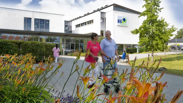 Auf dem Bild ist ein modernes, weißes Gebäude mit großen Fenstern zu sehen. Im Vordergrund wachsen bunte Blumen und grüne Sträucher. Zwei Personen, eine Frau in einem roten Shirt und ein Mann in einem karierten Hemd, gehen auf dem gepflasterten Weg, während im Hintergrund eine Person in rosa Kleidung zum Gebäude läuft. Ein Baum und eine Bank mit einer sitzenden Person vervollständigen die Szene.
