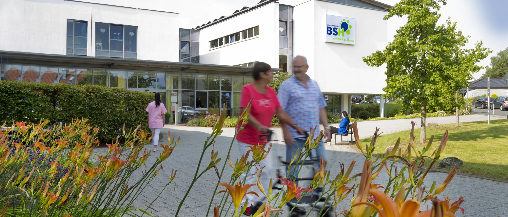 Leuchtend orangefarbene Blumen ragen in den Vordergrund des Bildes und bilden einen schönen Kontrast zu den hellen Gebäuden im Hintergrund. Zwei Menschen, ein Mann mit Rollator und eine Frau, gehen gemeinsam auf einem gepflasterten Weg. Im Hintergrund sie