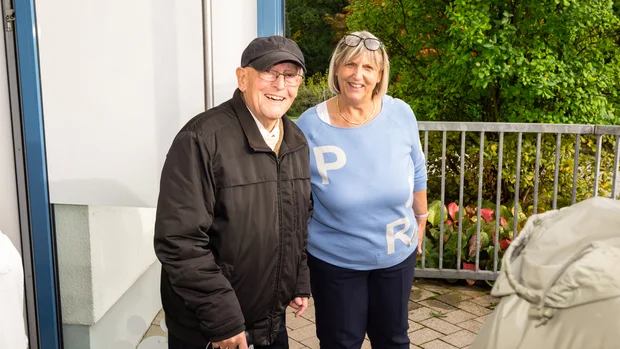 Ein älterer Mann mit Brille und schwarzer Jacke sowie einer schwarzen Schirmmütze steht lächelnd neben einer Frau mittleren Alters. Sie trägt einen hellblauen Pullover mit weißen Buchstaben 'P' und 'R'. Beide Personen wirken freundlich und stehen draußen vor einem Geländer mit grüner Bepflanzung im Hintergrund.