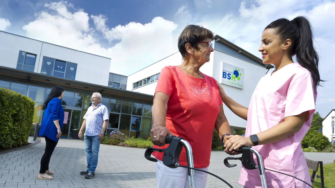 Auf dem Bild steht eine ältere Frau mit einem Rollator vor einem modernen, weißen Gebäude mit dem Schild „BSH – in Hagen zu Hause“. Eine junge Pflegekraft in rosa Kleidung legt ihr unterstützend eine Hand auf die Schulter, beide schauen sich freundlich an. Im Hintergrund unterhalten sich ein Mann in einem karierten Hemd und eine Frau in einer blauen Jacke vor dem verglasten Eingangsbereich des Gebäudes. Der Himmel ist blau mit weißen Wolken, und grüne Büsche und Blumen rahmen die Szene ein.