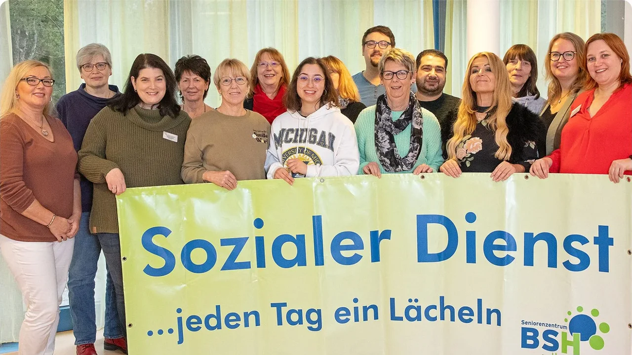 Eine Gruppe von Menschen steht gemeinsam in einem Raum und hält ein großes, hellgrünes Banner mit der Aufschrift „Sozialer Dienst … jeden Tag ein Lächeln“. Das Logo des Seniorenzentrums BSH ist in der unteren rechten Ecke des Banners zu sehen. Die Gruppe, bestehend aus Männern und Frauen, lächelt freundlich in die Kamera. Der Hintergrund zeigt große Fenster mit hellen Vorhängen, durch die Tageslicht einfällt.