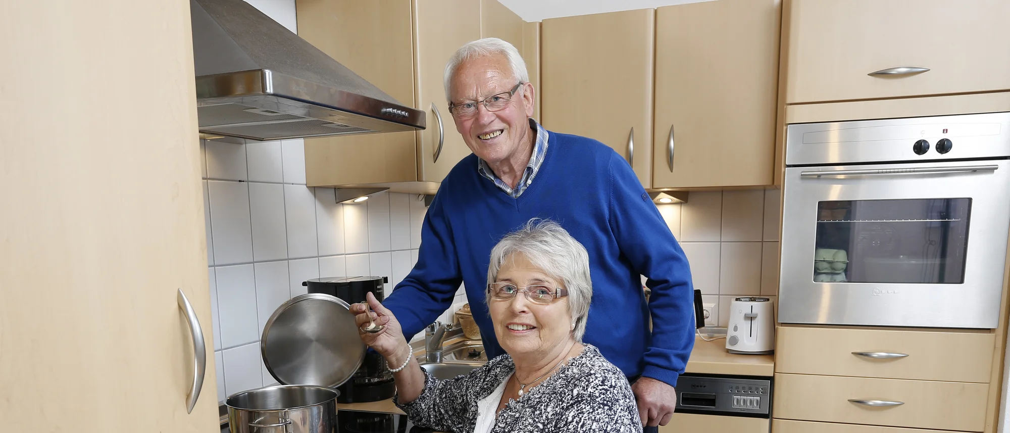 Ein älteres Paar ist in einer modernen Küche zu sehen. Die Frau sitzt in einem Rollstuhl und kocht, während der Mann hinter ihr steht und ihr hilft. Auf der Arbeitsfläche liegen ein Korb mit Äpfeln und Tomaten, was auf eine leckere Mahlzeit hindeutet.