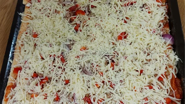 Ein großes Backblech ist mit einer selbstgemachten Pizza belegt. Auf der Pizza liegen rote Paprikastücke, Zwiebelringe und eine dicke Schicht geriebener Käse, der die Zutaten fast vollständig bedeckt. Die Pizza ist noch roh und bereit, in den Ofen geschoben zu werden.