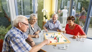 Vier ältere Menschen sitzen um einen Tisch und spielen ein Brettspiel. Sie scheinen Spaß zu haben, während sie konzentriert ihre Spielzüge machen. Der Hintergrund zeigt große Fenster mit Blick auf einen sonnigen Garten.