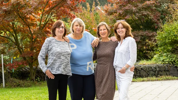 Vier Frauen unterschiedlichen Alters stehen lachend im Grünen vor buntem Herbstlaub. Sie haben ihre Arme umeinander gelegt und schauen fröhlich in die Kamera. Die Kleidung reicht von sportlich bis elegant, eine der Frauen trägt eine Brille. Die Szene vermittelt Zusammenhalt und Freundschaft in einer natürlichen Umgebung.