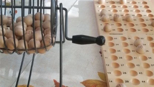 Eine Bingo-Trommel aus Metall, gefüllt mit kleinen Holz-Bingo-Kugeln, steht auf einem Tisch. Daneben liegt ein Holzbrett mit nummerierten Vertiefungen, in denen bereits einige Kugeln platziert wurden. Die Szene deutet auf ein Bingo-Spiel hin, das in einer gemütlichen Atmosphäre stattfindet, passend für gesellige Aktivitäten.