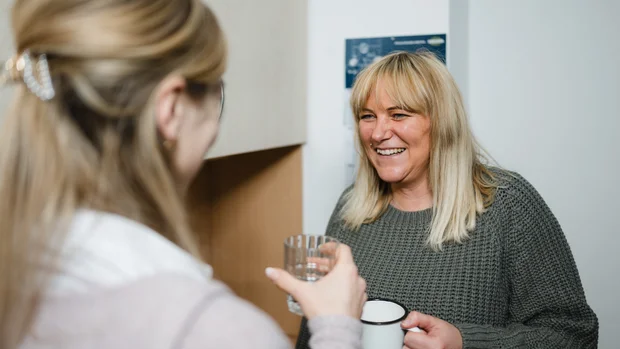 Das Bild zeigt zwei Personen in einem freundlichen Gespräch. Die Person im Vordergrund ist leicht unscharf und hat blondes Haar, das mit einer Haarspange zusammengehalten wird. Sie hält ein Wasserglas in der Hand. Die andere Person im Hintergrund trägt einen grünen Strickpullover und hat schulterlanges, blondes Haar mit Pony. Sie lacht herzlich und hält eine Tasse in der Hand. Der Hintergrund zeigt eine helle Wand mit einem aufgehängten Plan oder Poster.