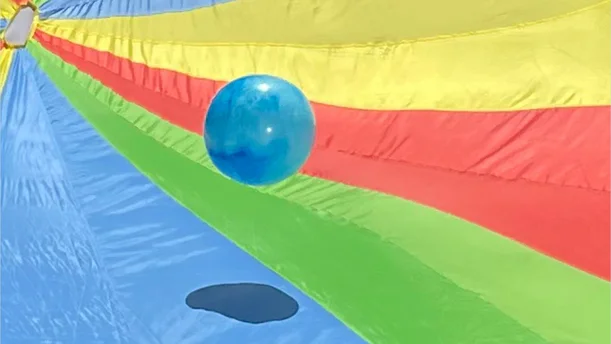 Eine blaue Kugel schwebt über einem bunten Schwungtuch mit Streifen in Blau, Grün, Gelb und Rot. Das Tuch scheint in Bewegung zu sein, wodurch die Kugel leicht angehoben wird. Die kräftigen Farben des Tuchs und die schwebende Kugel erzeugen eine spielerische und fröhliche Atmosphäre.
