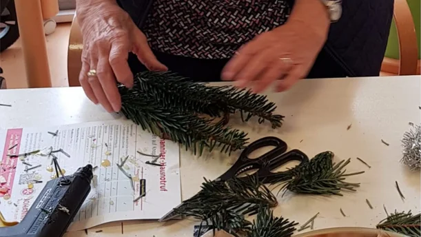 Eine Person arbeitet an einem Tisch mit Tannenzweigen und Bastelmaterialien. Auf dem Tisch liegen eine Schere, eine Heißklebepistole und ein Blatt Papier mit Bastelanweisungen. Die Szene deutet darauf hin, dass ein kreatives Projekt, wie das Gestalten eines Adventskranzes oder einer Weihnachtsdekoration, in Arbeit ist.