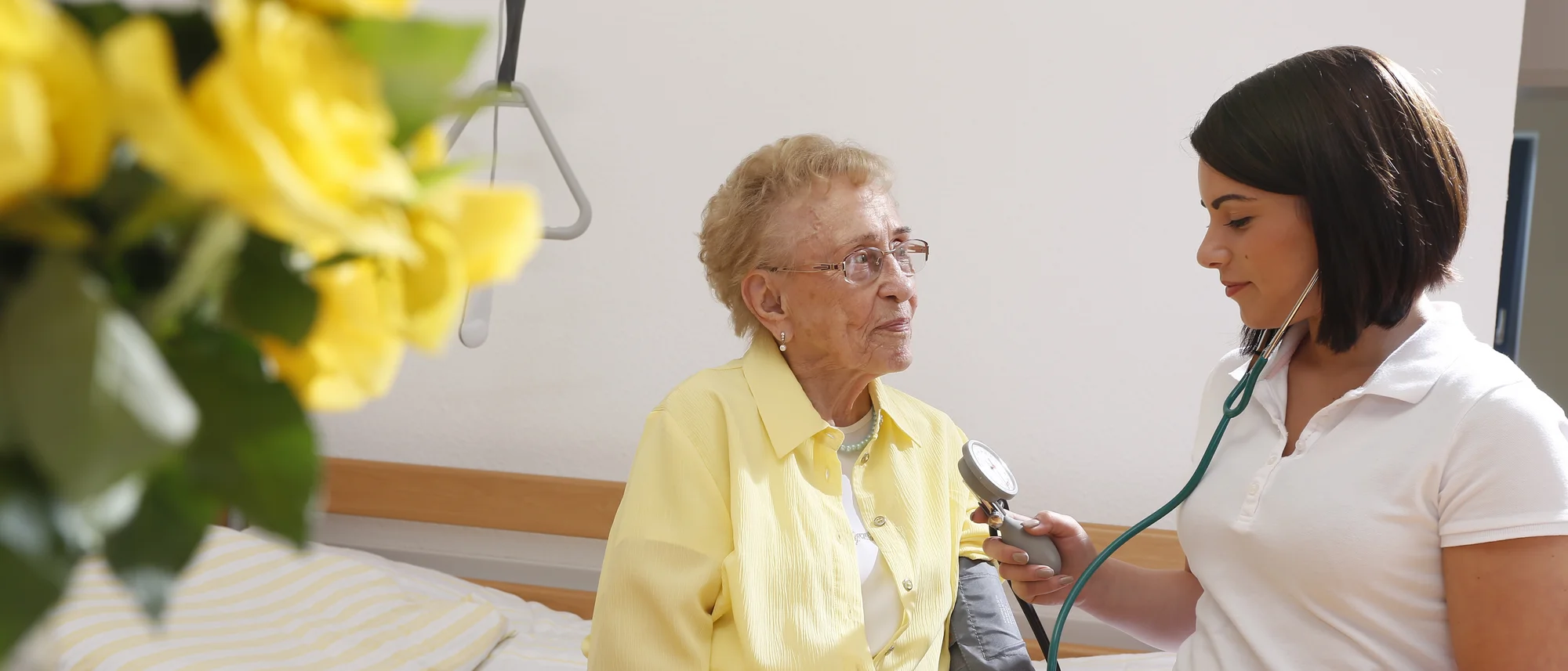 Das Bild zeigt eine ältere Frau in einer gelben Bluse, die auf einem Bett sitzt und von einer jungen Frau in weißer Pflegekleidung medizinisch betreut wird. Die junge Frau misst mit einem Blutdruckmessgerät den Blutdruck der Seniorin und hört dabei konzentriert zu. Im Vordergrund sind gelbe Blumen zu sehen, die eine warme und freundliche Atmosphäre schaffen. Der Hintergrund ist schlicht und lenkt den Fokus auf die Interaktion zwischen den beiden Personen.