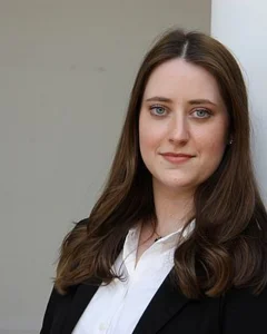 Das Bild zeigt eine Person mit langen, dunklen Haaren, die sich leicht gewellt über die Schultern legen. Sie trägt eine weiße Bluse und darüber ein schwarzes Jackett. Die Person lehnt entspannt an einer weißen Säule und blickt freundlich in die Kamera. Der Hintergrund ist neutral und schlicht gehalten.