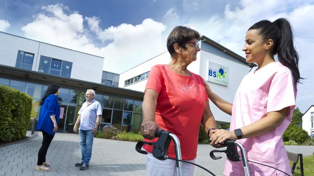 Zu sehen sind eine ältere Frau im roten T-Shirt und eine jüngere Frau in pinker Arbeitskleidung. Sie wirken freundlich und lächeln sich an, während sie vor einem modernen Gebäude stehen. Der Hintergrund zeigt grüne Büsche, bunte Blumen und das Logo des Seniorenheims "BSH".