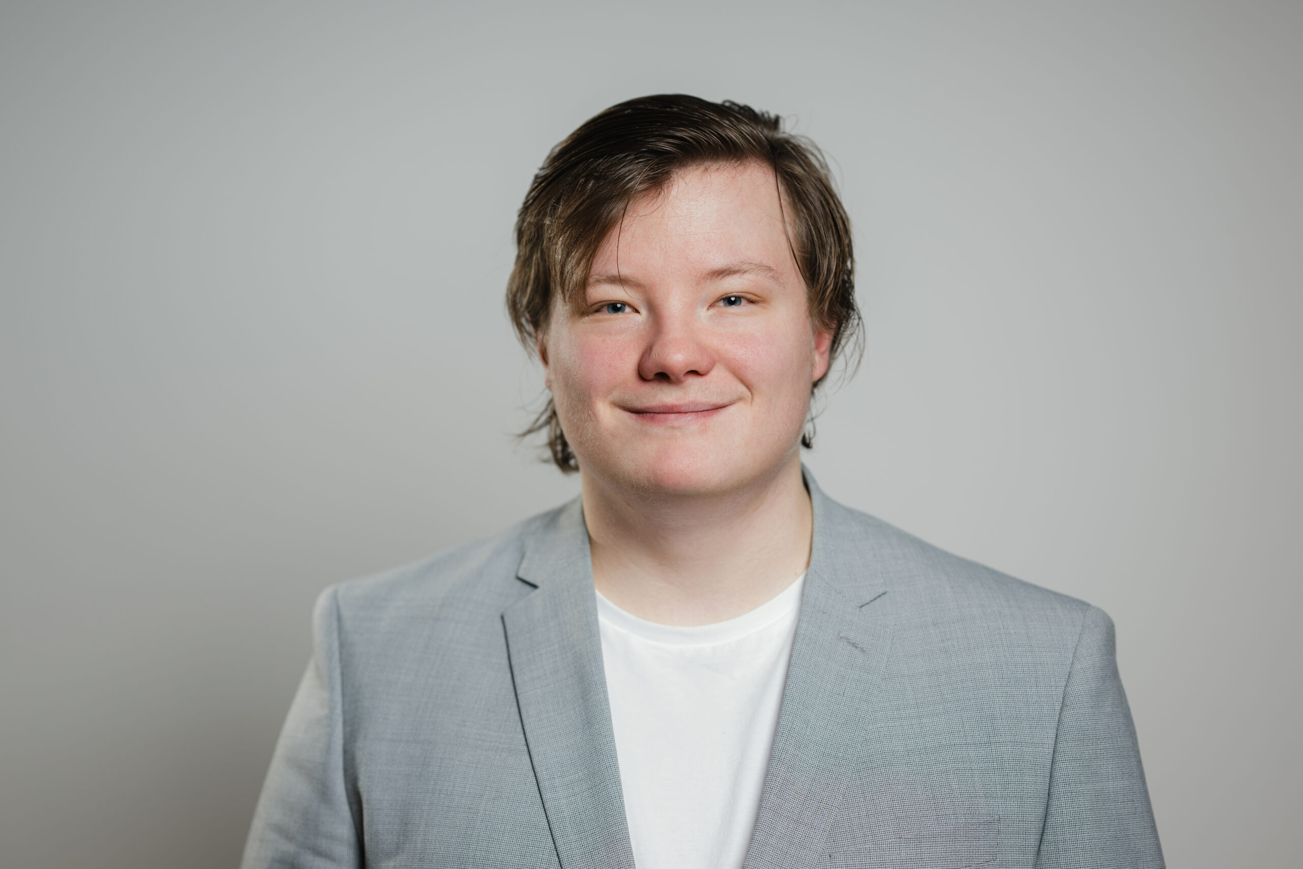 Das Bild zeigt eine Person mit mittellangem, leicht gewelltem Haar, die freundlich in die Kamera lächelt. Sie trägt ein hellgraues Sakko über einem weißen T-Shirt. Der Hintergrund ist neutral grau gehalten, sodass das Porträt im Mittelpunkt steht.