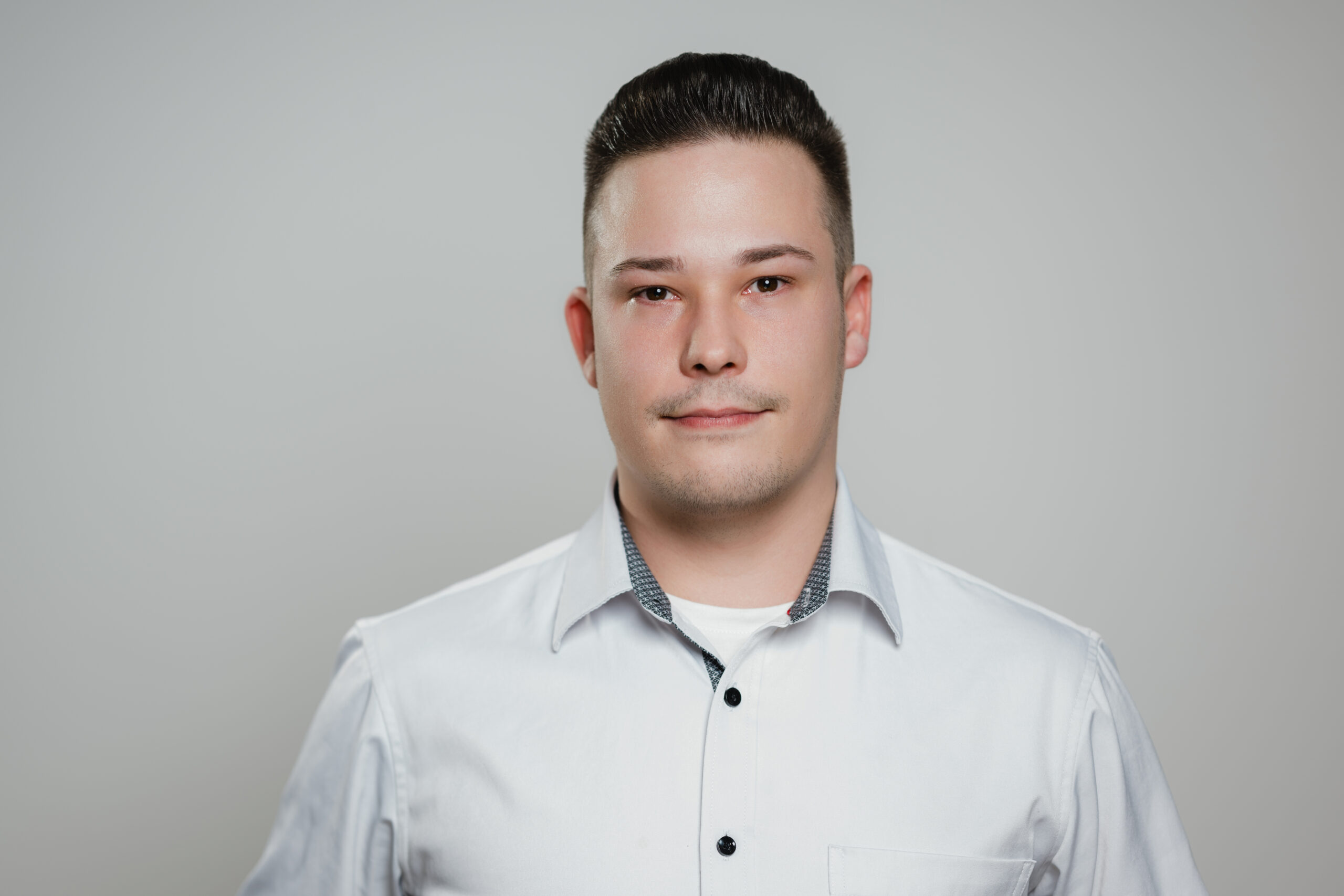 Das Bild zeigt eine Person mit kurzem, dunklem Haar, die ein weißes Hemd mit einem dunklen Kragen trägt. Die Person schaut direkt in die Kamera mit einem neutralen Gesichtsausdruck. Der Hintergrund ist schlicht und grau, was das Porträt betont.
